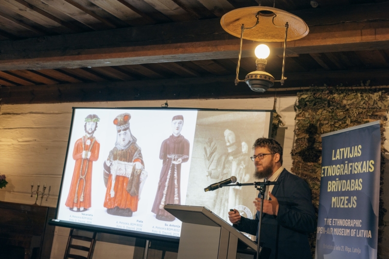Referents uzstājas konferencē, fonā eksrāns ar svēto attēliem