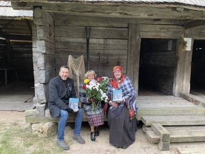 Trīs cilvēki ar ziediem un jauno Lībiešu kultūras mantojuma katalogu sēž uz klēts lieveņa.