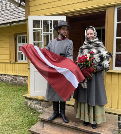 Pie Jaunsaimniecības stāv jaunieši tautastērpos ar Latvijas karogu un sarkanbaltsarkanām rozēm.