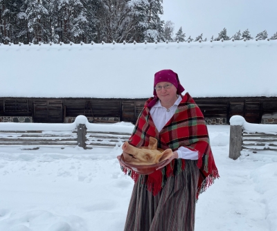 Sieviete tautastērpā nes cūkas galvu māla bļodā pāri Kurzemes sētas pagalmam.