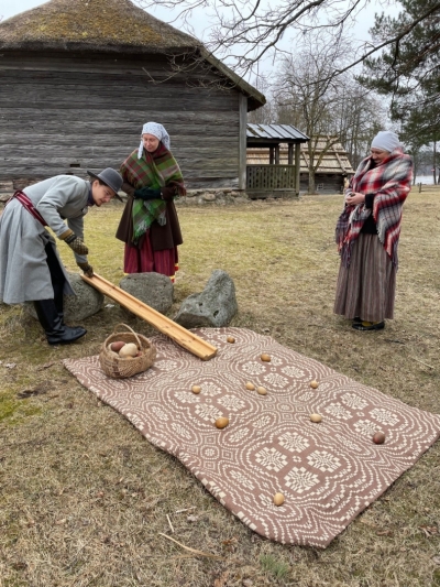 Trīs cilvēki tautastērpos demonstrē Lieldienu tradīciju – olu ripināšanu. 