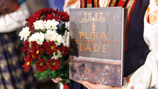 Sieviete tautastērpā tur rokās jauno izdevumu “Pūra lāde” un sarkanbaltsarkanu ziedu pušķi. 
