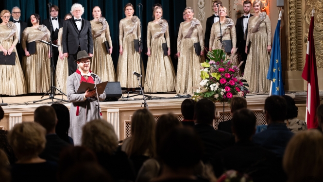 Svinību atklāšana: uz skatuves koris ar diriģentu; muzeja pārstāvis uzrunā viesus pie Latvijas un ES karogiem. 