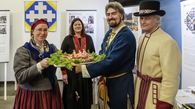 Četri cilvēki tautastērpos tur svētku kliņģeri ar degošām svecēm skaitļa “100” formā. 