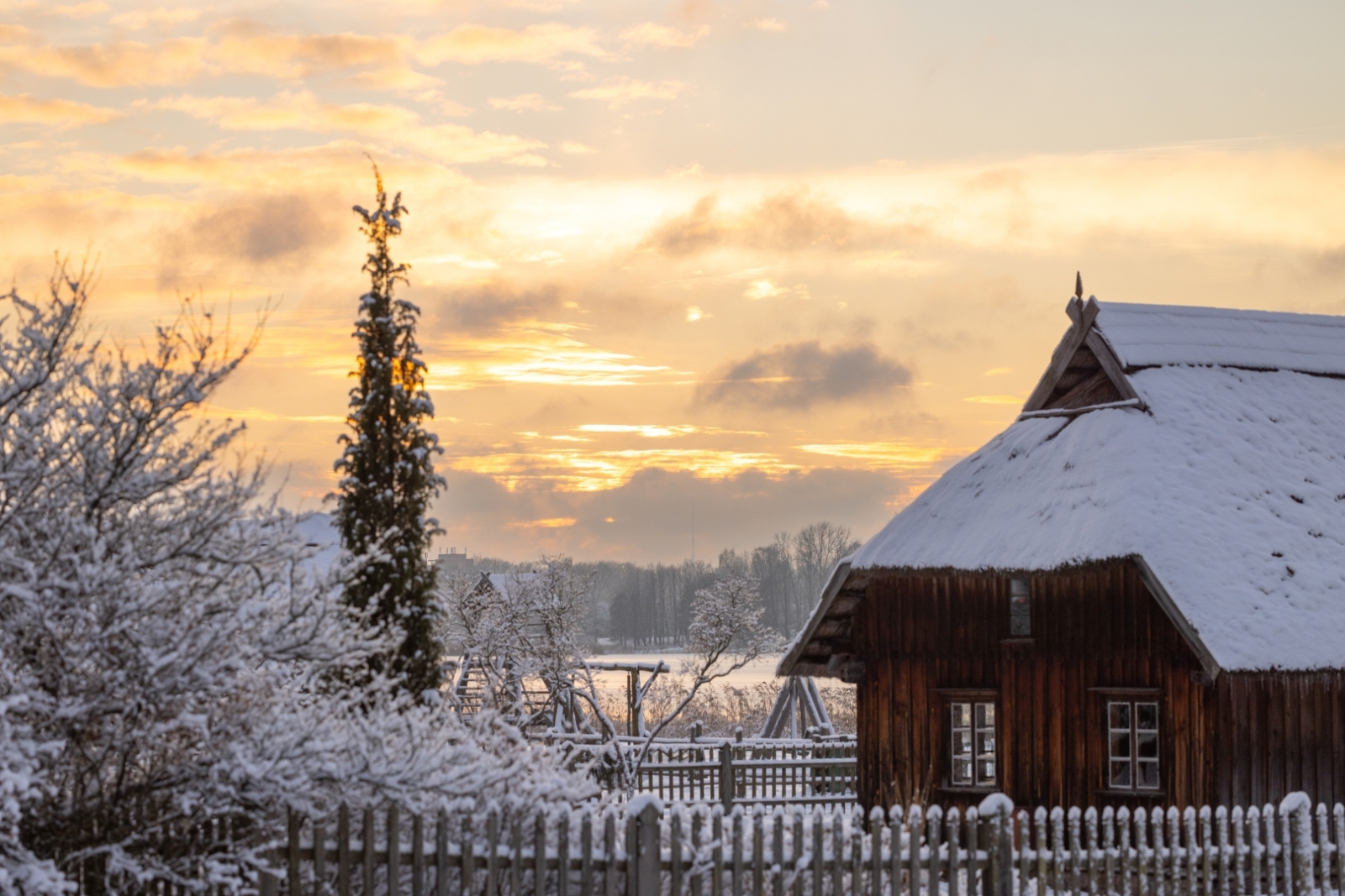 Latvijas Etnogrāfiskā brīvdabas muzeja ziemas ainava ar apsnigušām vēsturiskajām koka ēkām.