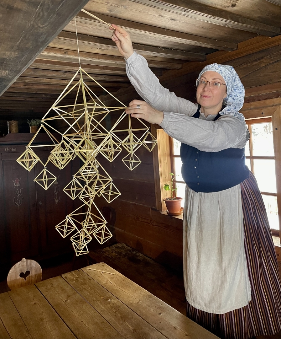 Sieviete tautastērpā rotā telpu, karot pie koka griestu sijām tradicionālu latviešu puzuru.