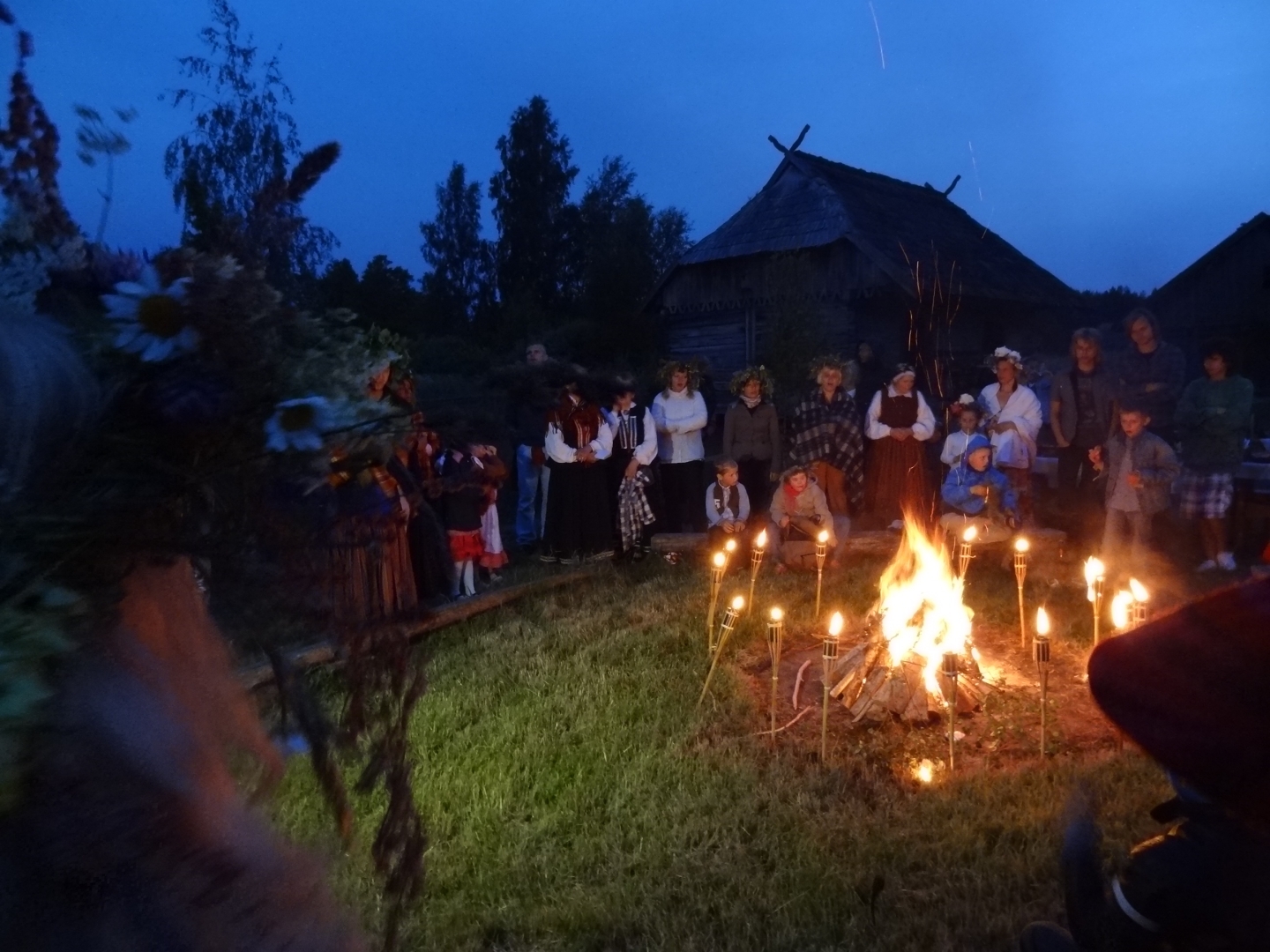 Jāņu ugunskurs "Vītolnieku" sētā, ap kuru iedegtas lāpas un stāv svinētāji.