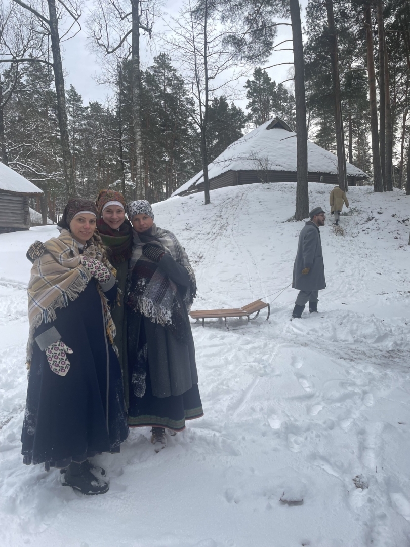 Trīs meitenes priecājas par ziemas priekiem; fonā puisis velk ragavas