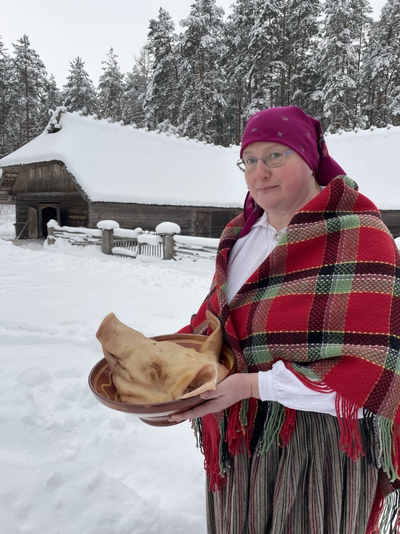 Sieviete tautastērpā nes pāri sniegotam lauku pagalmam lielu bļodu, kurā ir cūkas galva