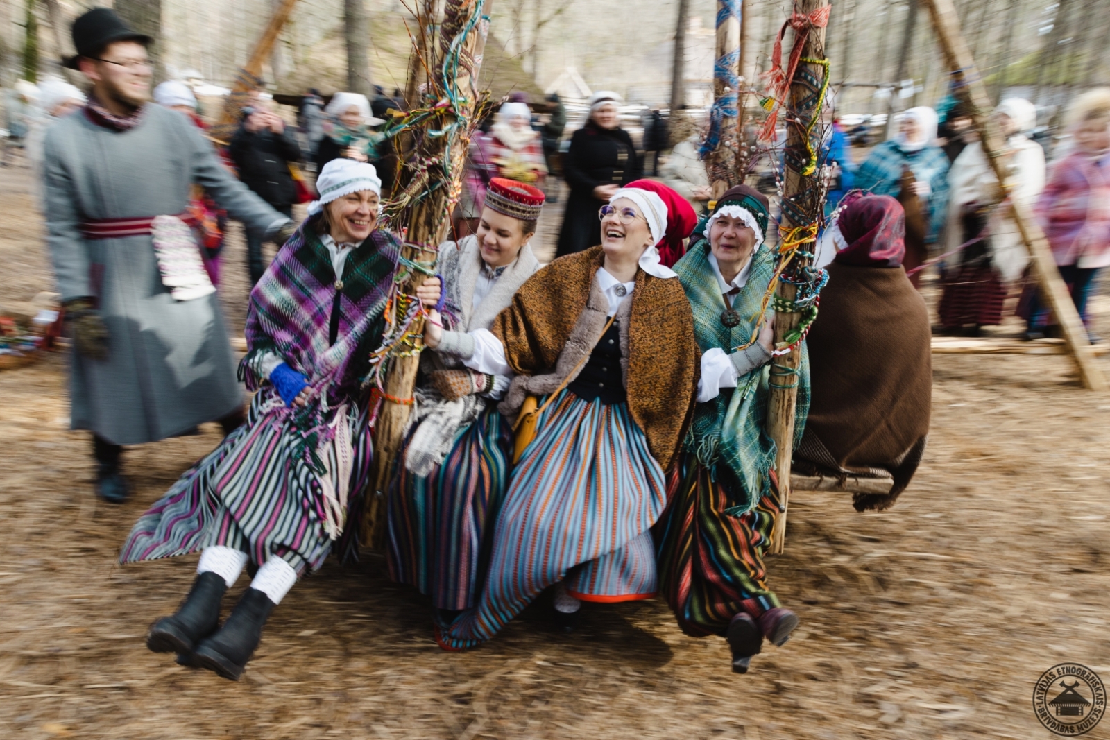 Sievietes krāšņos tautastērpos lielajās Lieldienu šūpolēs  šūpo puisis tautas tērpā