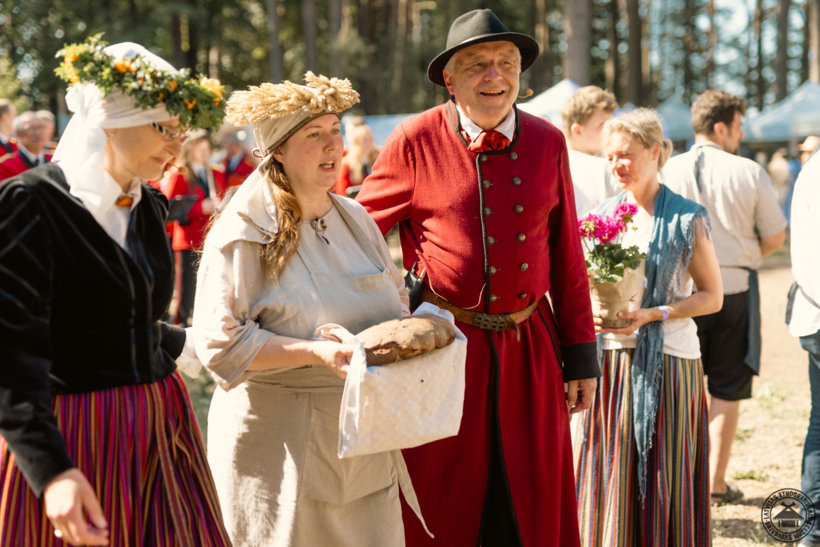 Tirgus kultūras programmas vadītāji kopā ar maizes cepēju, kurai galvā vārpu vainags un rokās svaigs maizes kukulītis. 