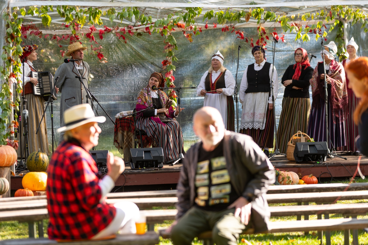 Skats uz skatuvi ar folkloras kopu, kamēr priekšplānā divi vīrieši sarunājas