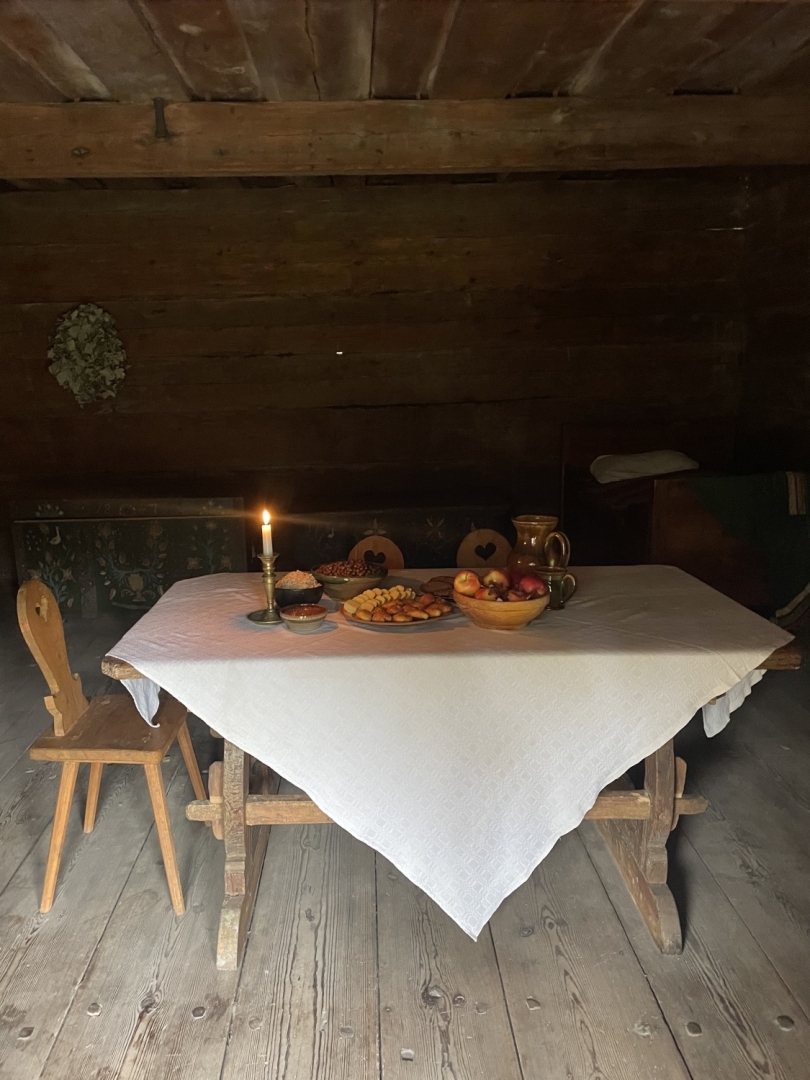 Veļu mielasta galds klētiņā, ko izgaismo viena degoša svece, radot noslēpumainu un mierīgu noskaņu.