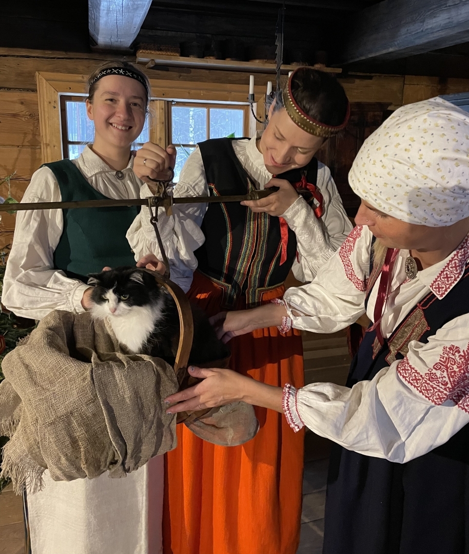 Meitas veic senu zīlēšanas rituālu, sverot kaķi uz vecmodīgiem svariem senatnīgā istabā