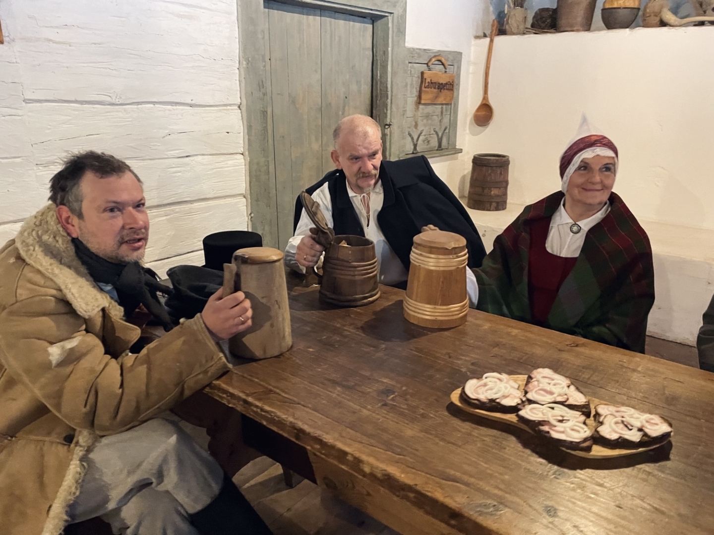 Svētku viesi krogā sēž pie gariem koka galdiem, malkojot alu no tradicionāliem kausiemSvētku viesi krogā sēž pie gariem koka galdiem, malkojot alu no tradicionāliem kausiem