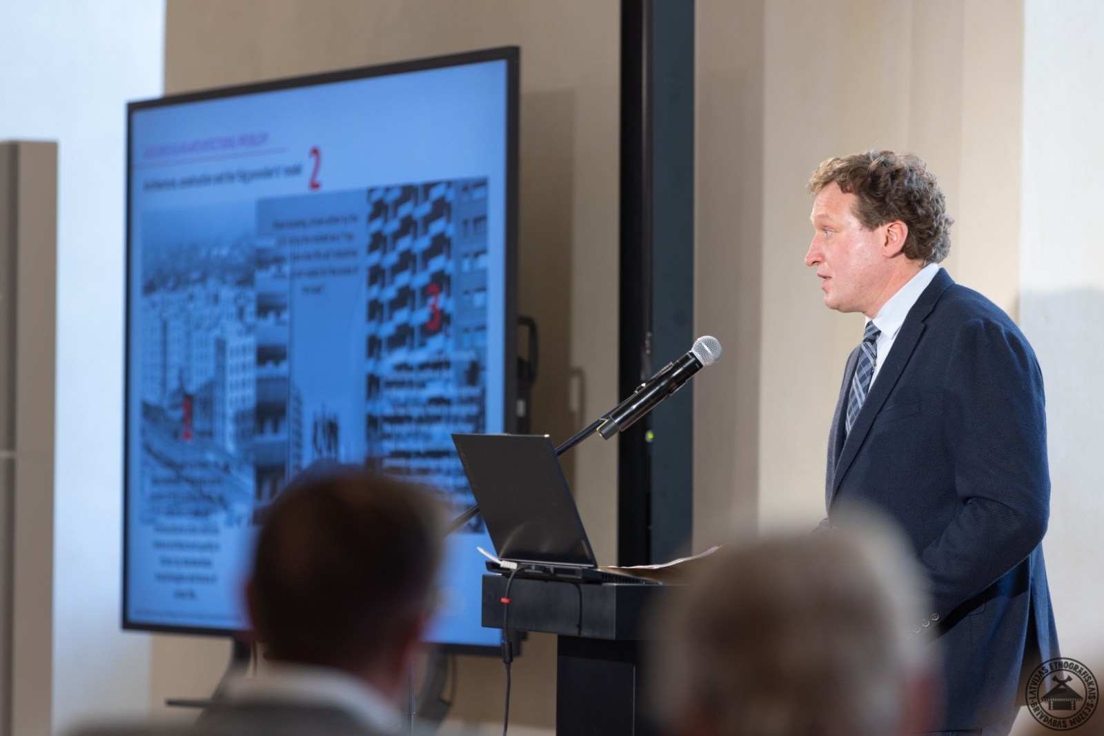 Lektors prezentē referātu, ekrānā redzams modernās augstceltnes attēls. 
