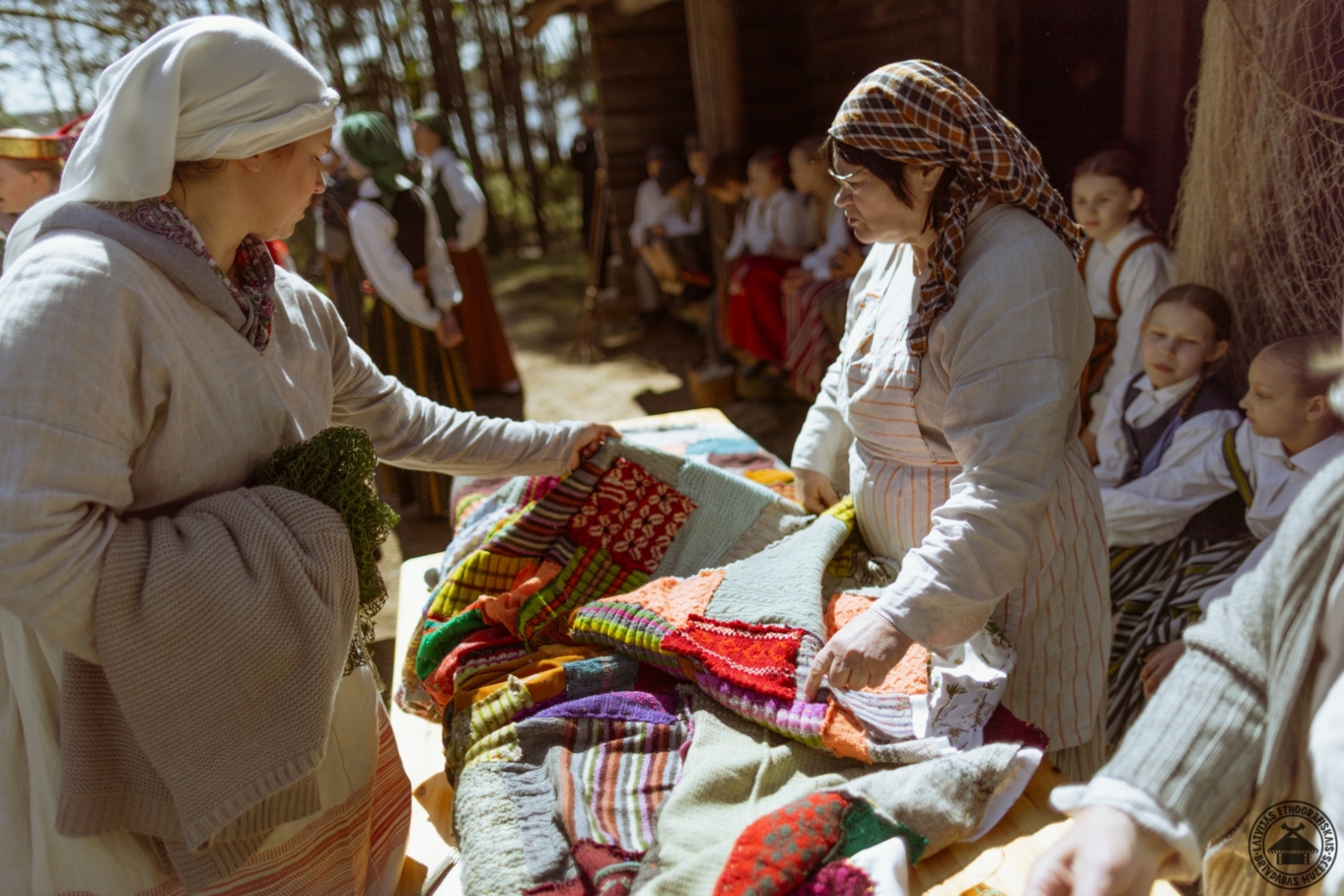 Sievietes tautastērpos kopīgi cilā, apskata un pēta dažādus košus, krāsainus un rakstainus plecu lakatus.