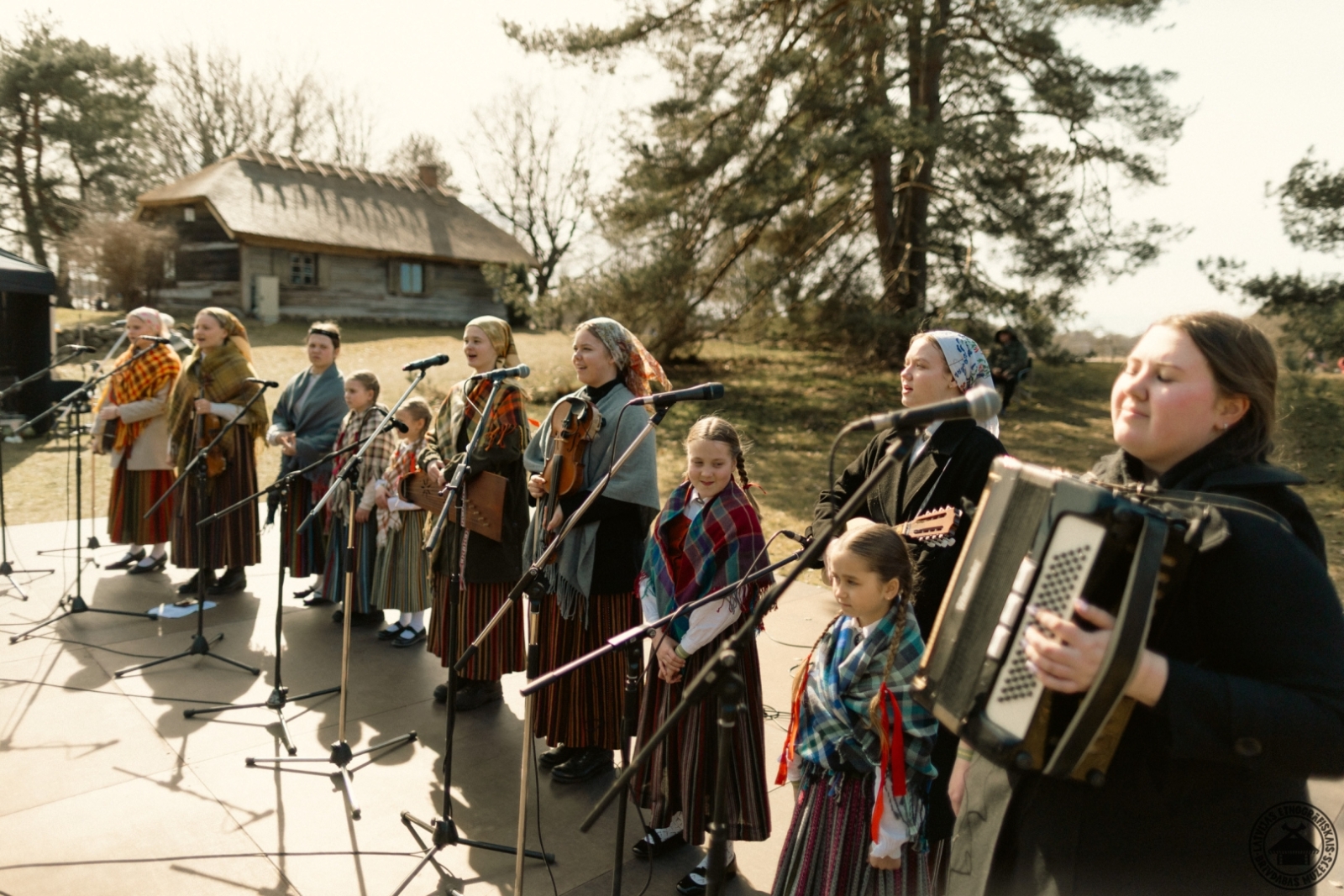 Folkloras kopa tautastērpos dzied Lieldienu dziesmas uz skatuves pie mikrofoniem.