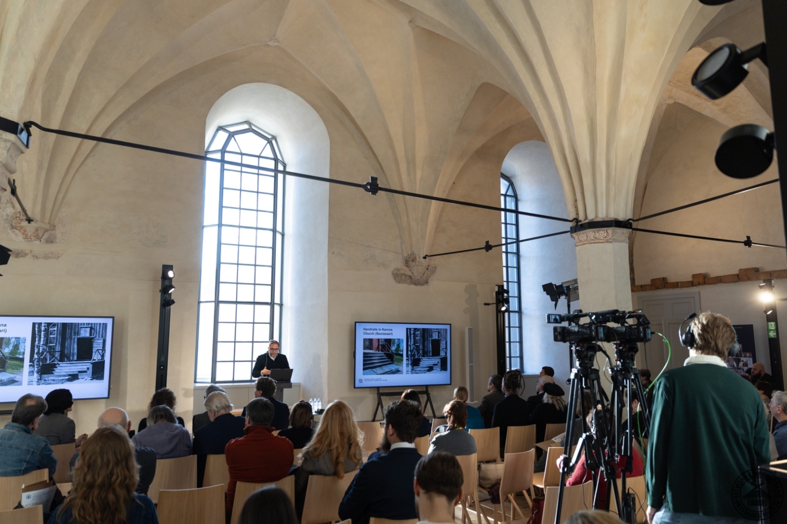 Konferences norise: priekšplānā videokamera, kas filmē pasākumu. 