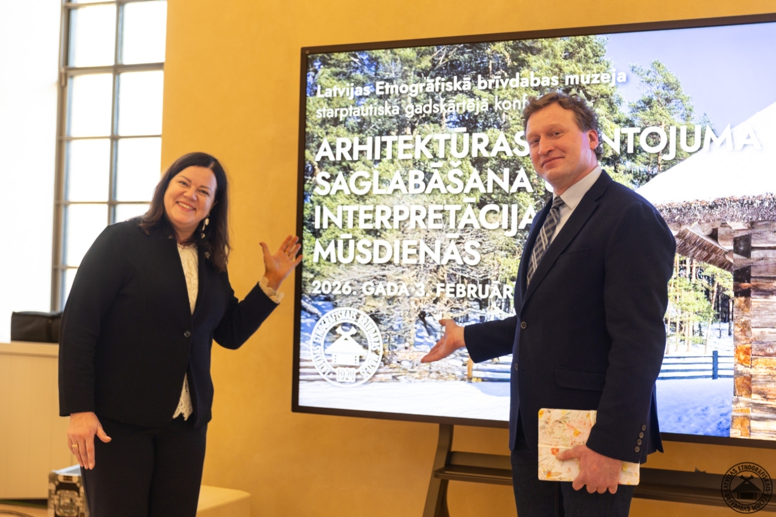 Muzeja direktore un direktora vietnieks stāv pie prezentācijas ekrāna konferencē par arhitektūras mantojumu. 