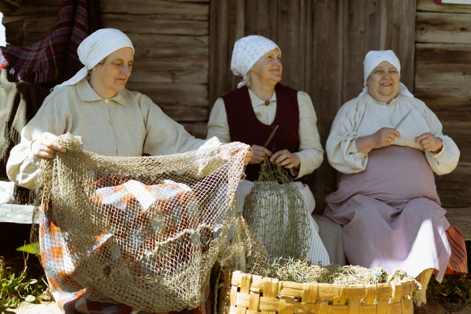 Trīs sievietes tautastērpos kopīgi cilā un ar rokām lāpa liela izmēra zvejas tīklu.
