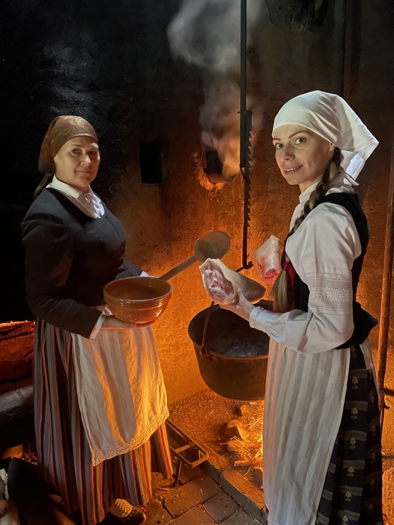 Divas sievietes stāv senā virtuvē pie pavārda, kurā karājas liels katls; vienai rokā ir gaļas gabals, otrai – koka bļodas.