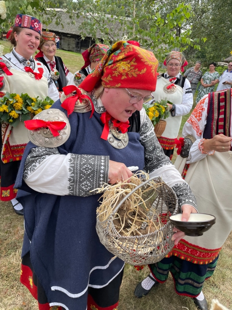 Kāzu rituāla norise, kur priekšplānā redzama sieviete krāšņā Rucavas tautastērpā ar villaini.