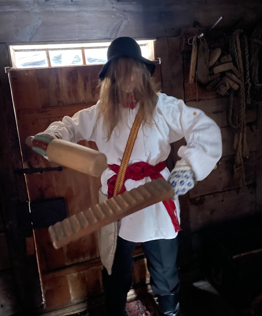 Meitene pilnā maskā un rokās viņa tur tradicionālos rīkus – veļas rulli un veļas vāli.