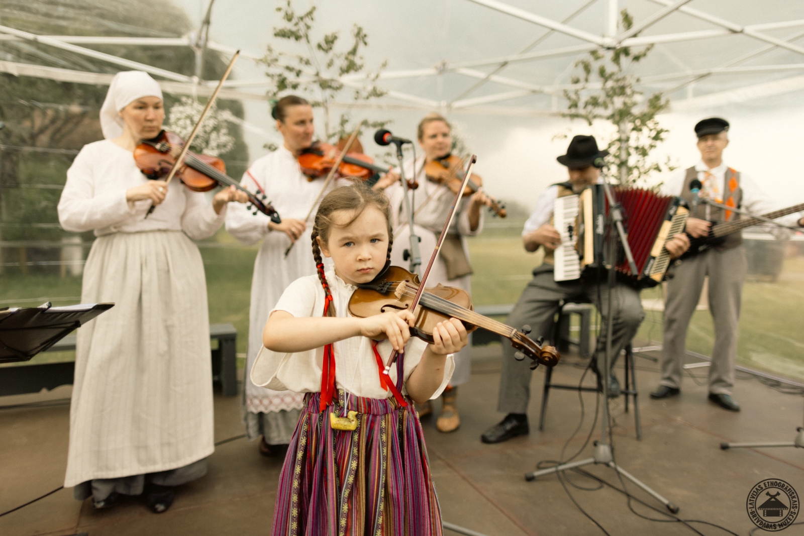 Folkloras grupas muzicēšana; priekšplānā maza meitenīte centīgi spēlē vijoli. 