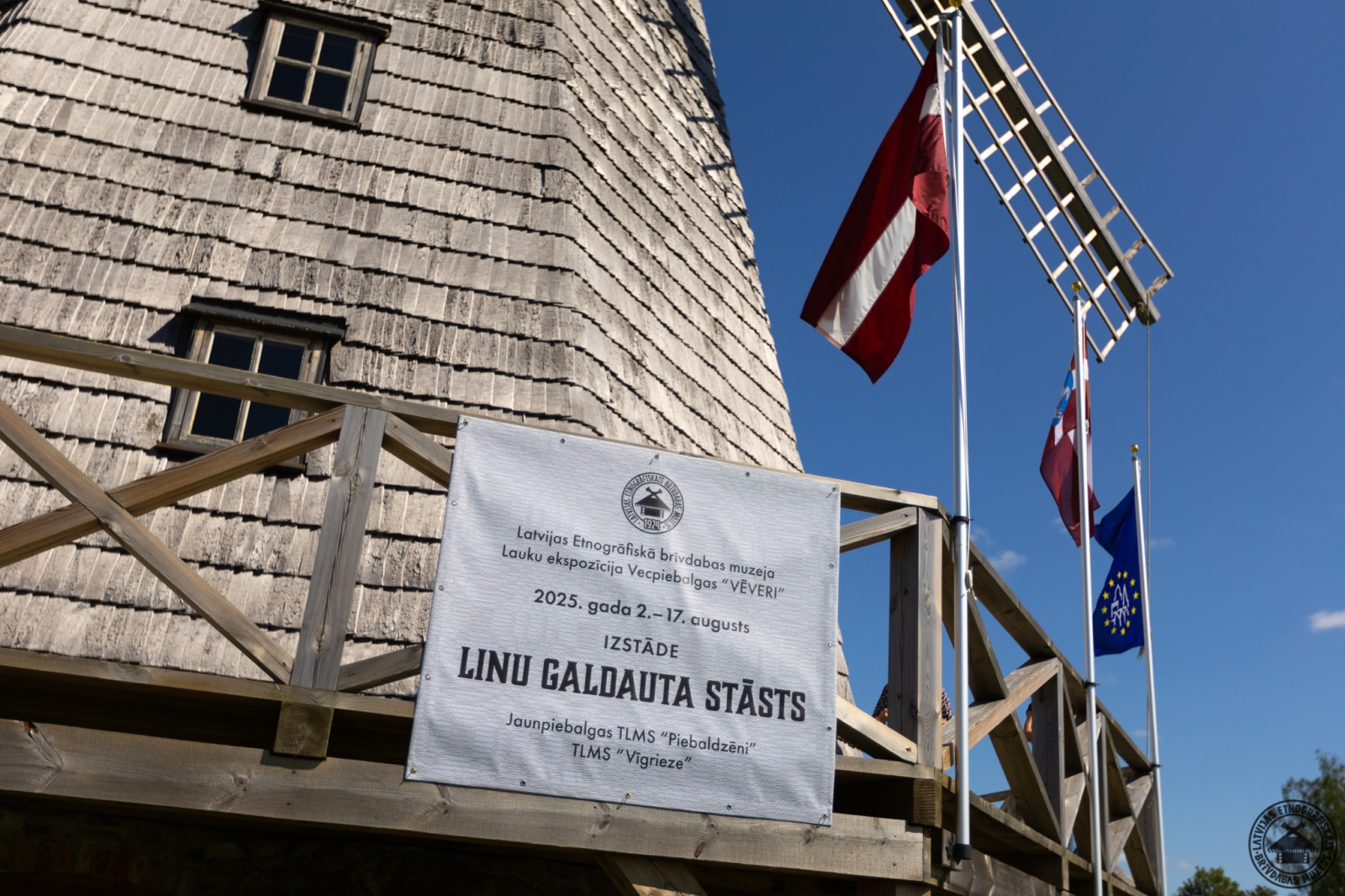 Vēveru vējdzirnavas ar afišu par izstādi “Linu galdautu stāsts”  