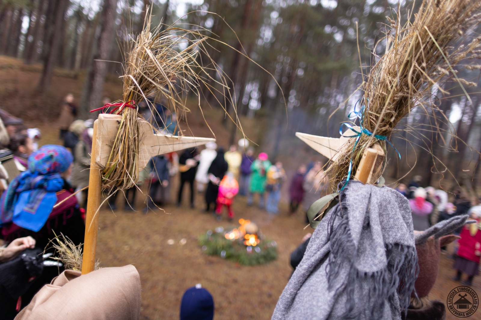 Cilvēki sildās ap ugunskuru; priekšplānā dalībnieki Putnu maskās. 
