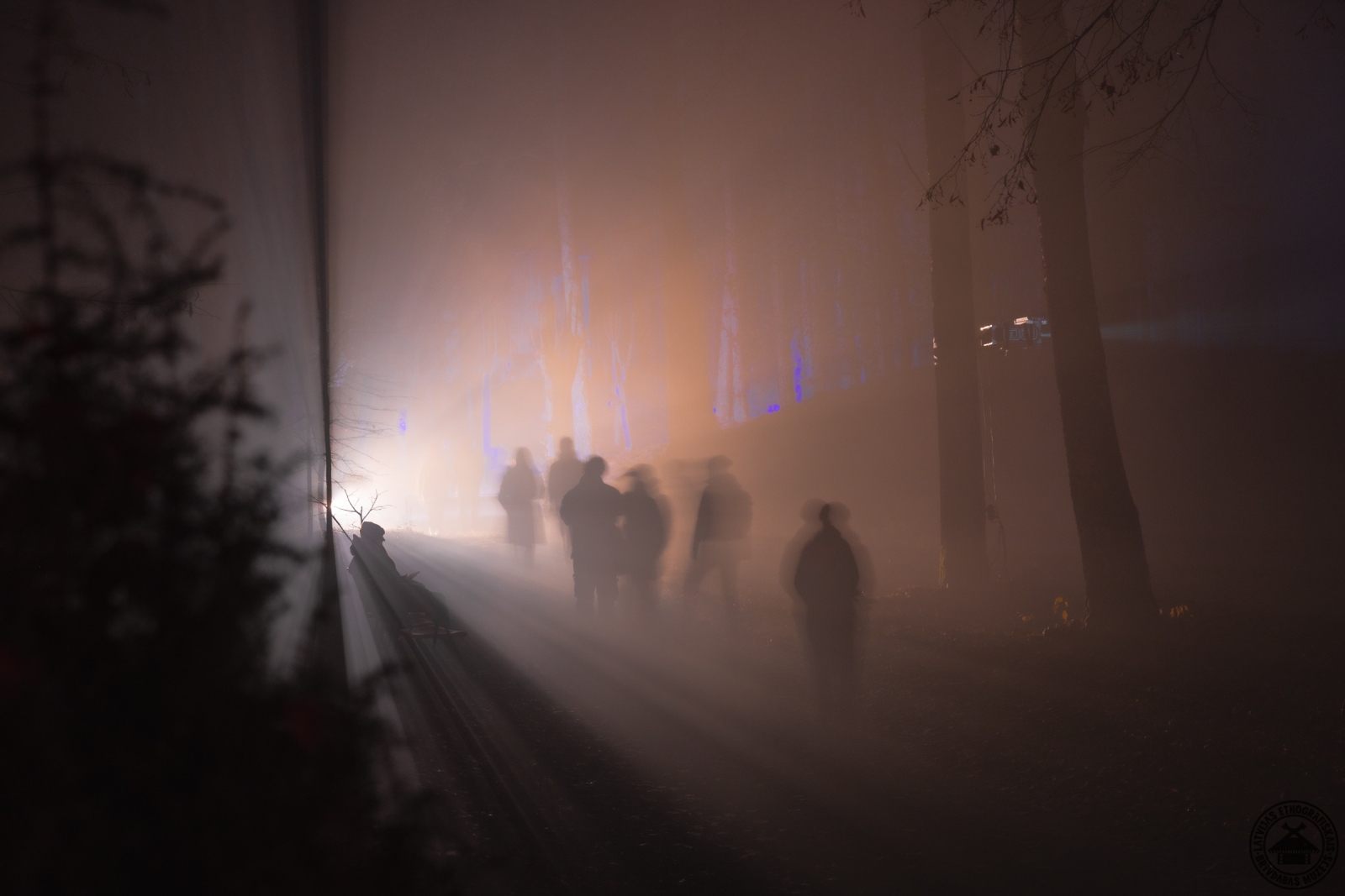 Cilvēku silueti biezā miglā un tumsā, radot maģisku un sirreālu viņsaules gaisotni. 