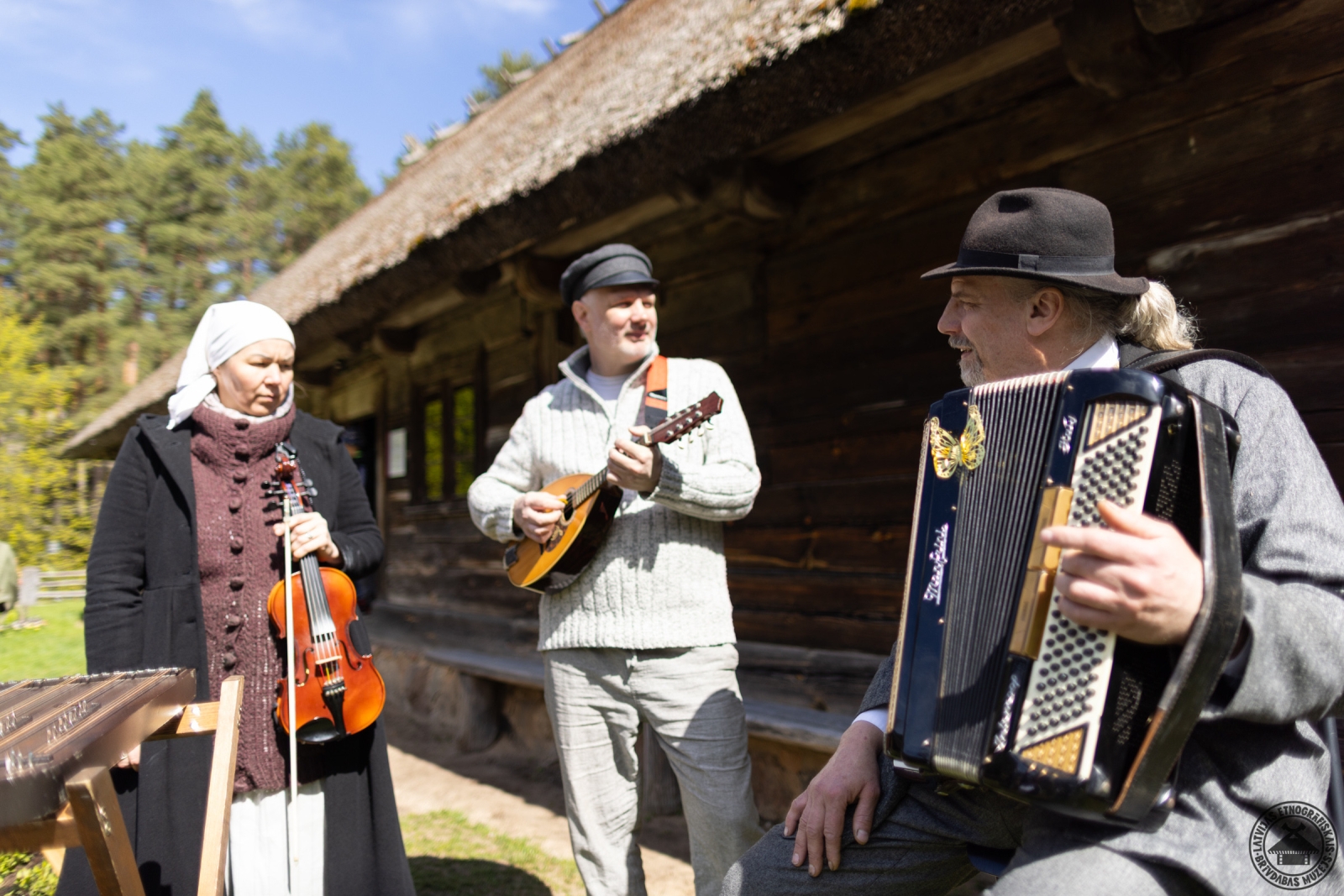 Muzikanti tautastērpos ar akordeonu, mandolīnu un vijoli pie Kurzemes dzīvojamās mājas. 