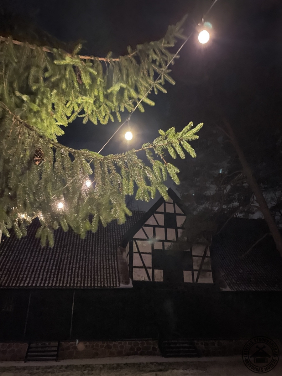 Izgaismoti Vidzemes sētas ēku jumti zem mākoņainām nakts debesīm sniegā. 