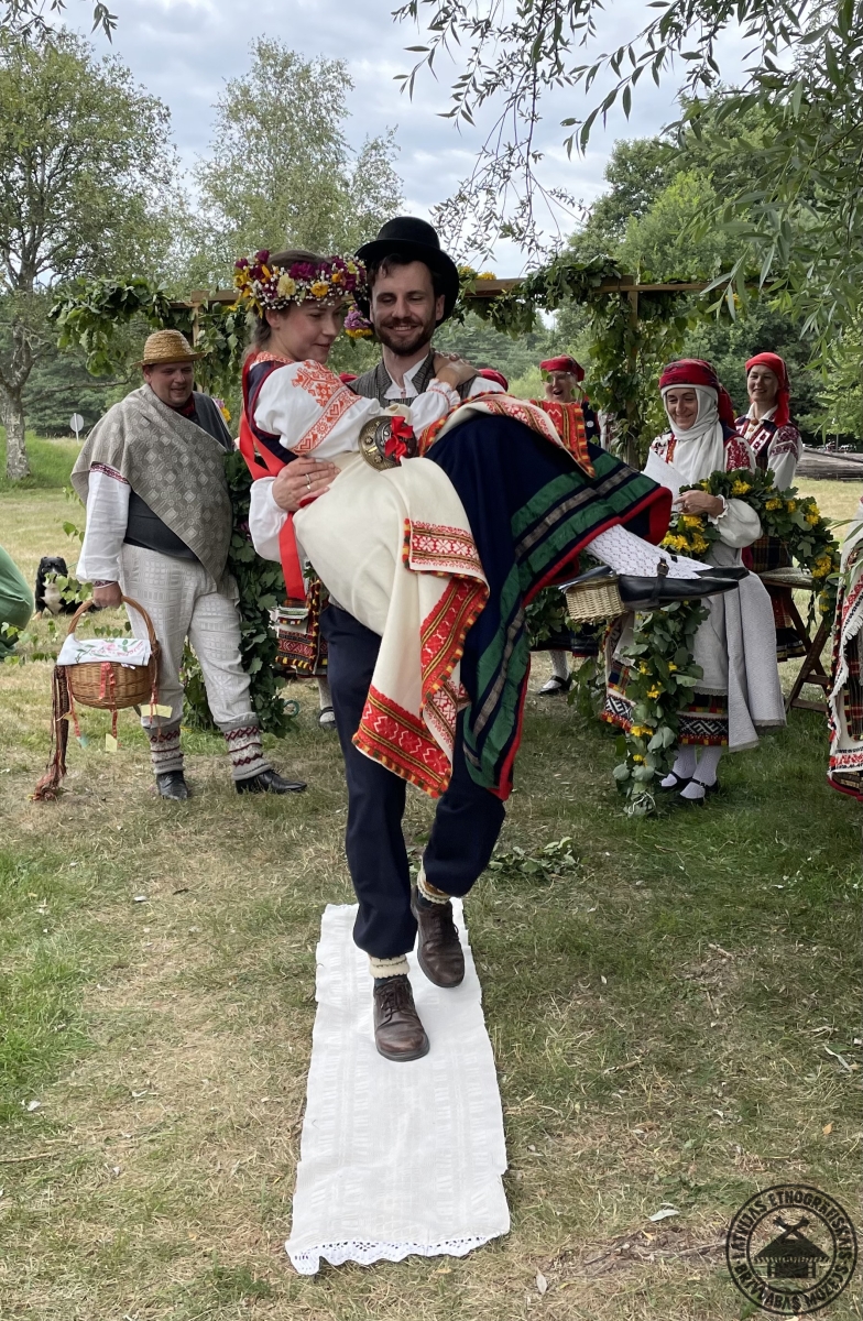 Līgavainis nes līgavu uz rokām pāri dvielim, zem kura paslēpts šķīvis zīlēšanas rituālam. 