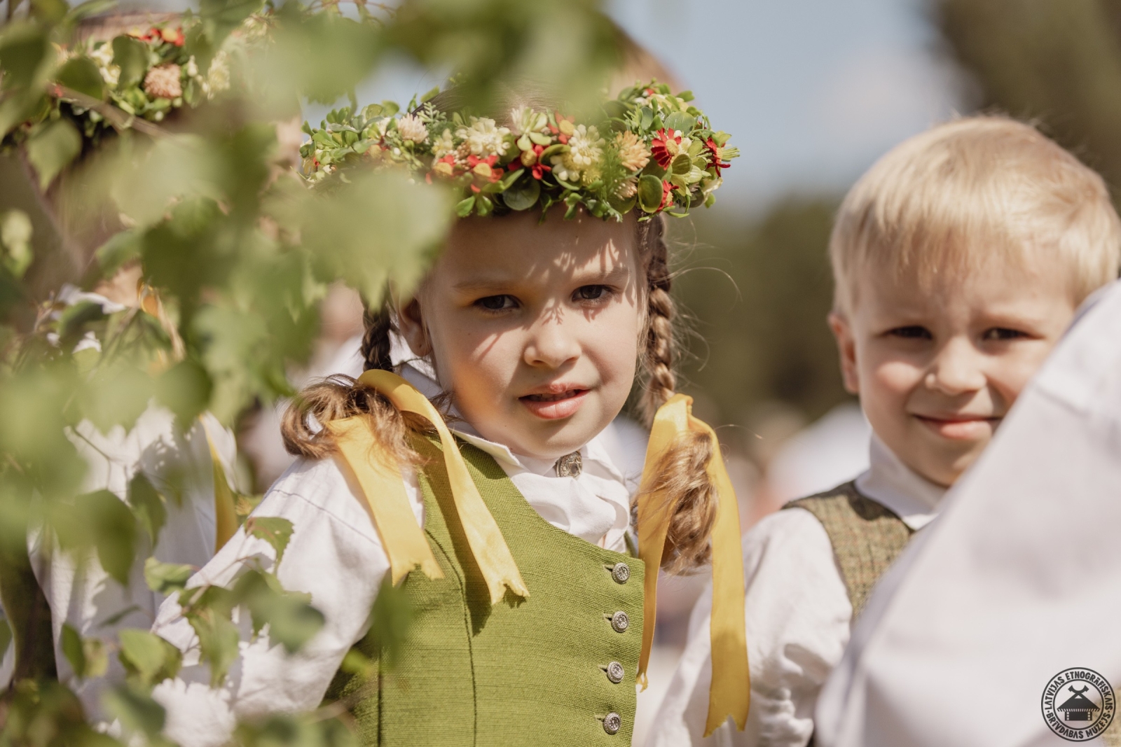 Divi mazi bērni tautastērpos; meitenēm galvā skaisti pļavas ziedu vainadziņi. 