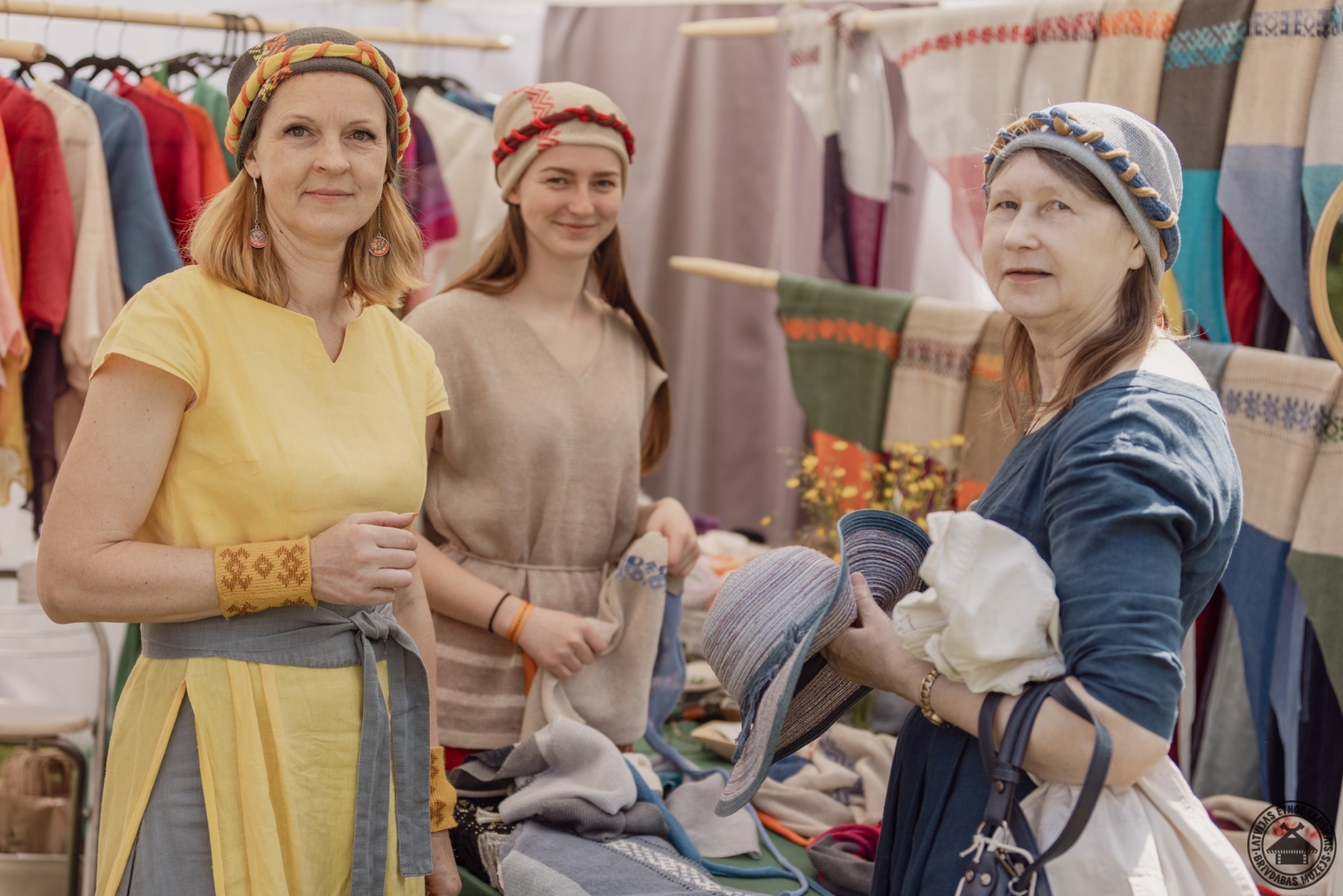 Rokdarbu stends ar adītām jakām un cepurēm; centrā smaidīgs pārdevējs. 