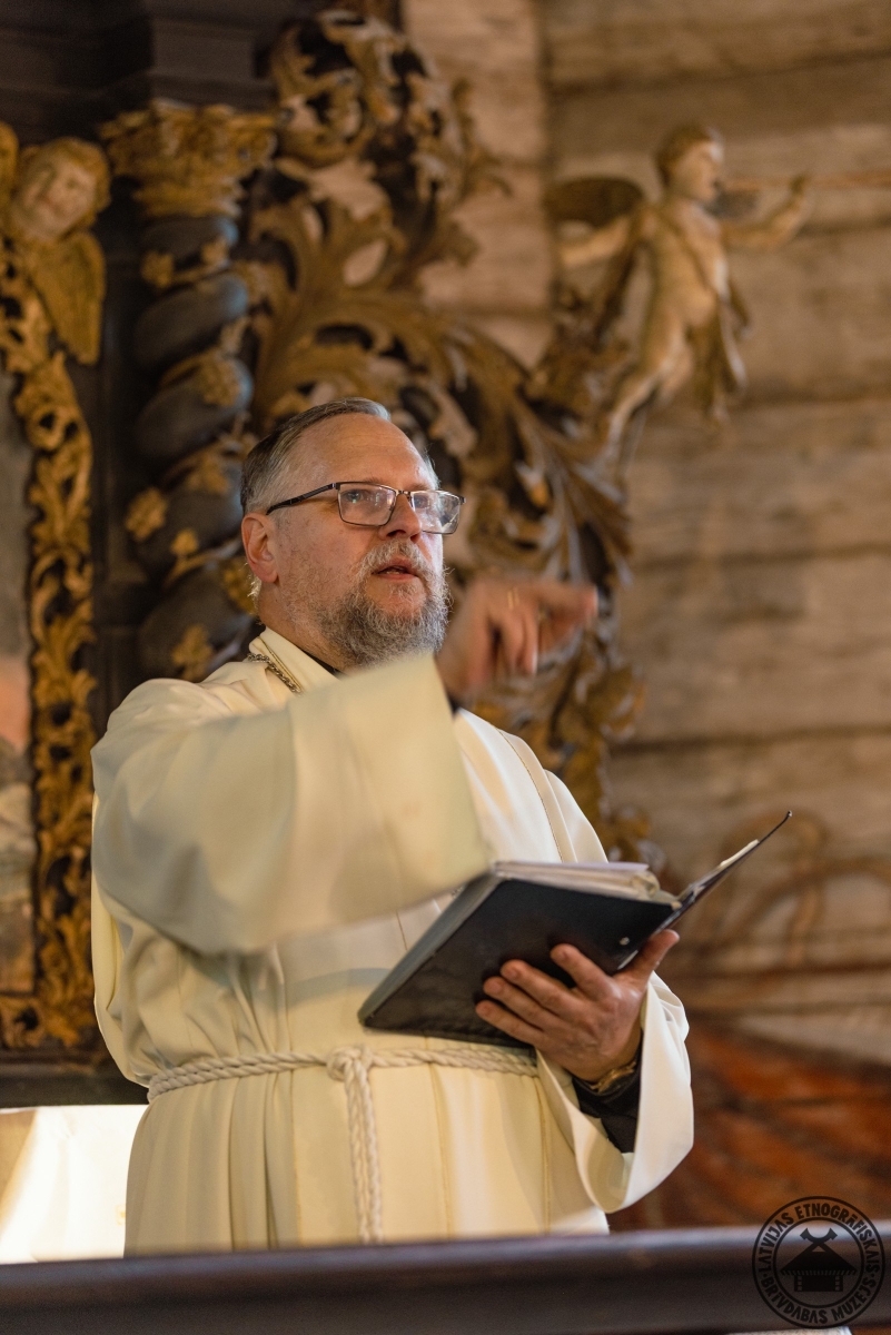 Mācītājs liturģiskajā tērpā ar Bībeli rokās vada svētku dievkalpojumu. 