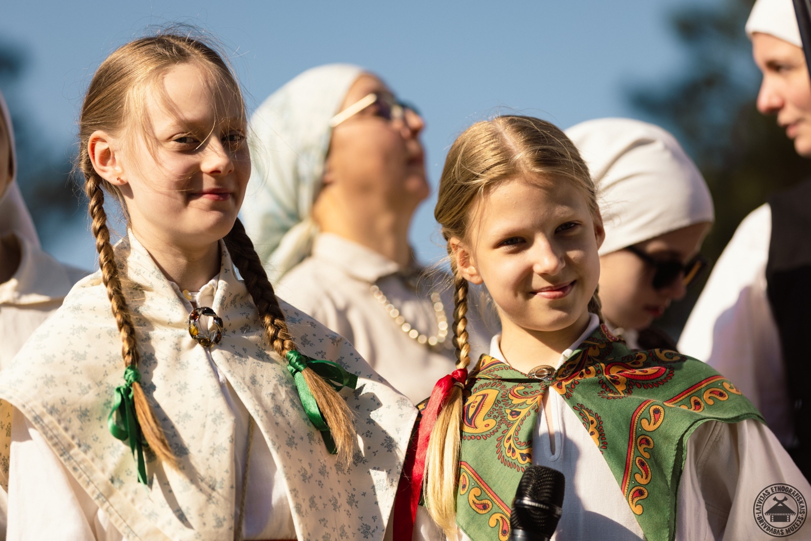 Divas meitenes ar garām bizēm un tautastērpos priecīgi smaida fotogrāfam. 