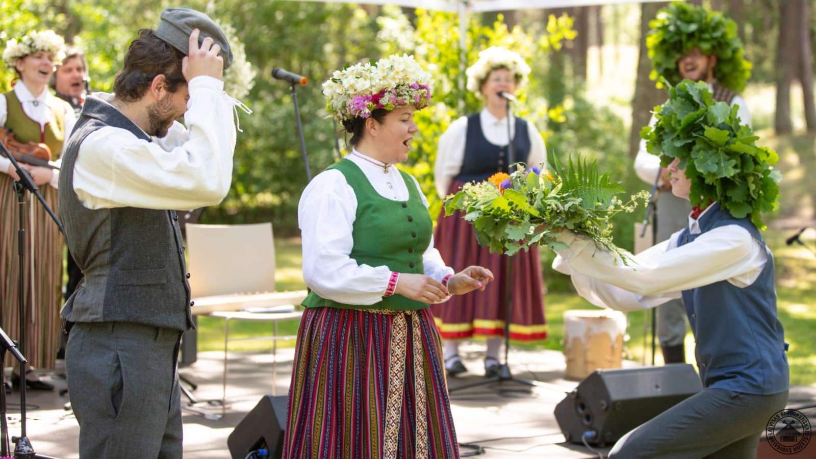 Muzeja direktorei tautastērpā un divos vainagos svinīgi tiek pasniegts kupls jāņuzāļu pušķis. 