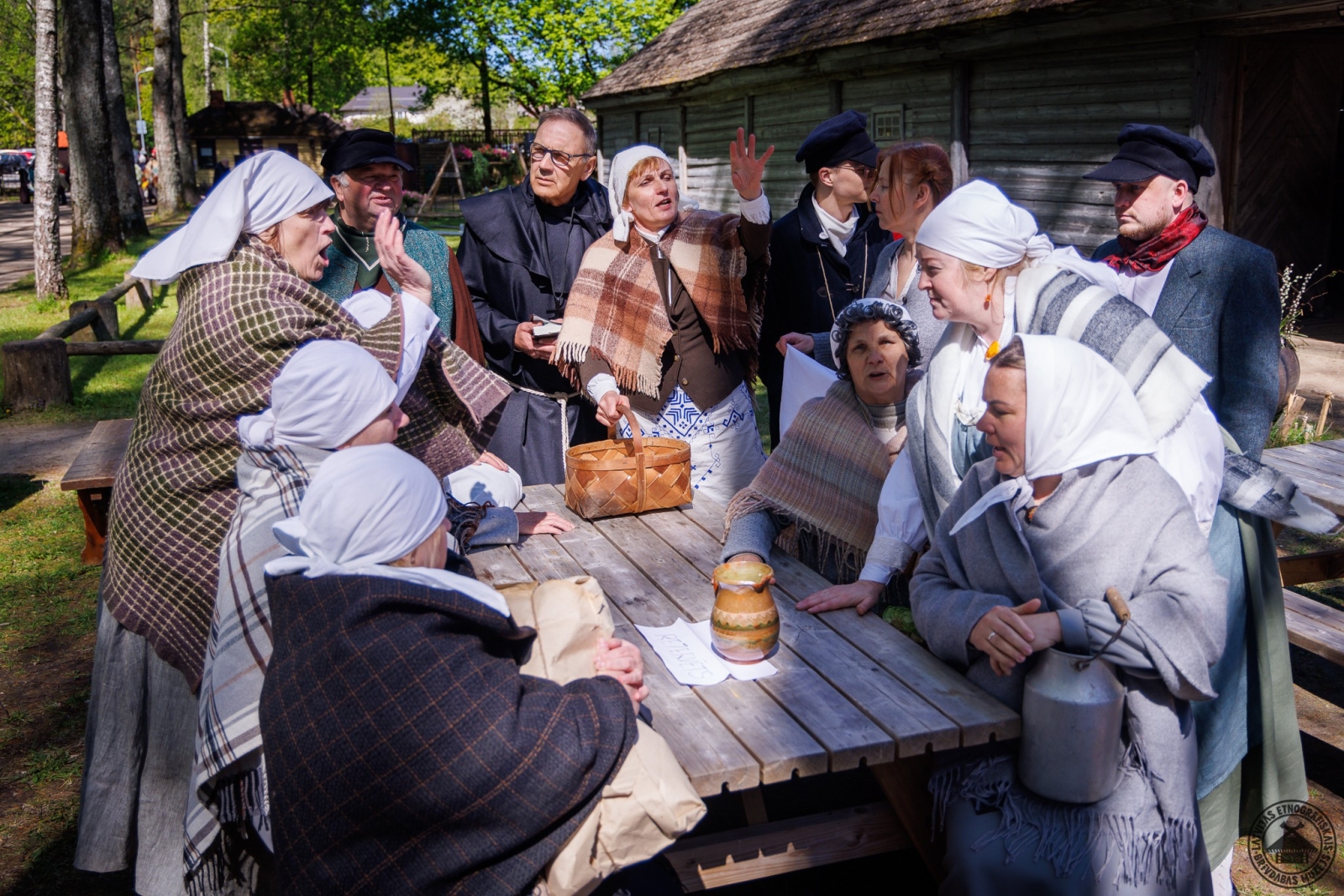 Liels pulks cilvēku senlaiku apģērbos pie Priedes kroga sasēduši un sastājušies ap galdu. 