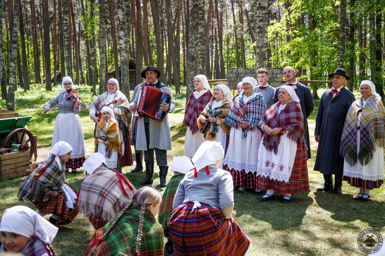 Folkloras kopa dzied; bērni priekšplānā iet rotaļā, nometušies tupus. 