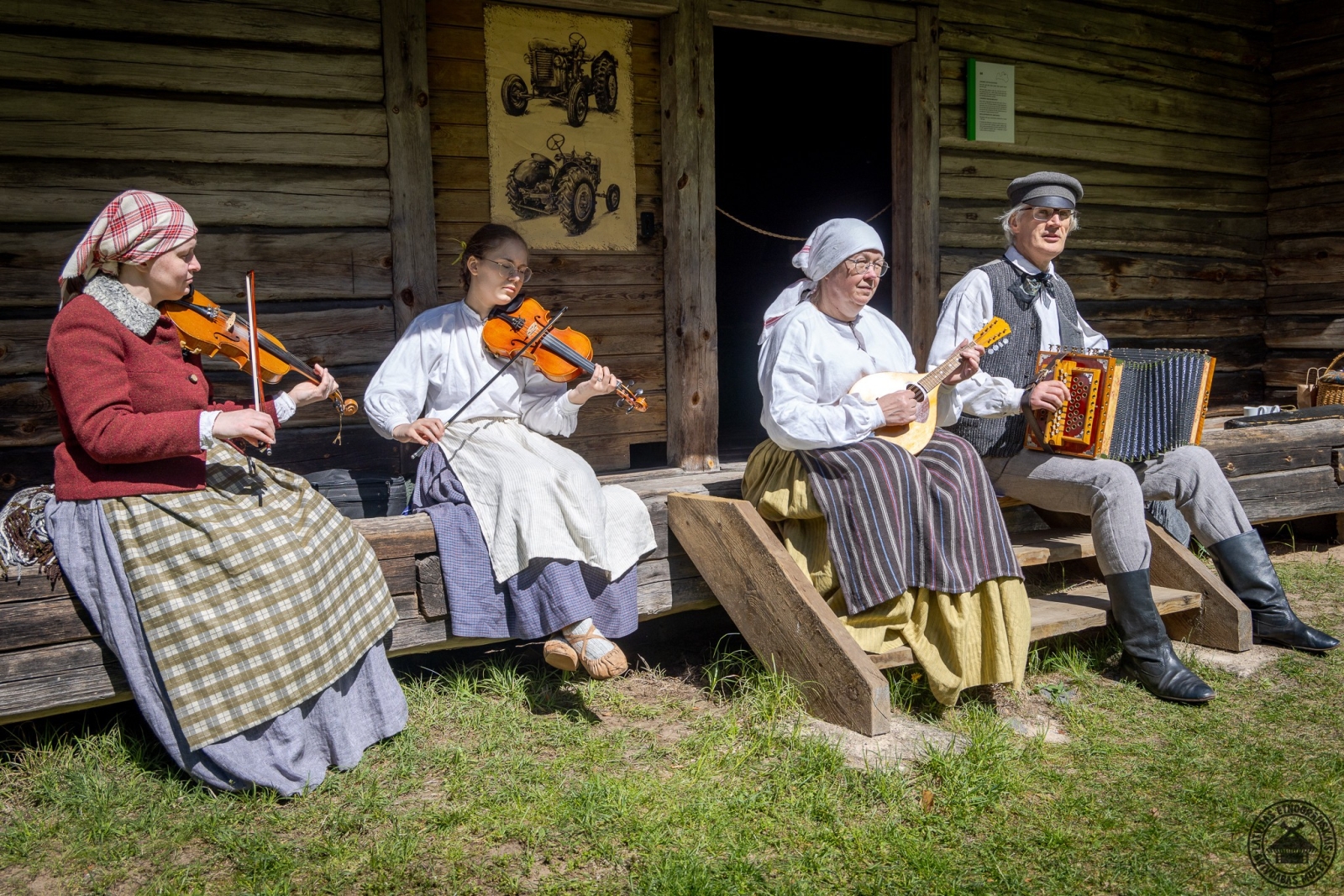 Uz Jaunsaimniecības klēts lieveņa sēž mūziķi, spēlējot vijoles, mandolīnu un akordeonu. 