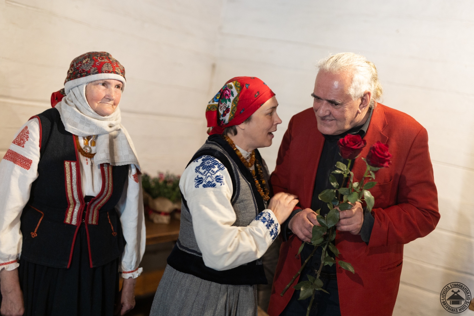 Divas sievietes krāšņos Rucavas tautastērpos sveic mākslinieku ar sarkanām rozēm izstādes atklāšanā. 