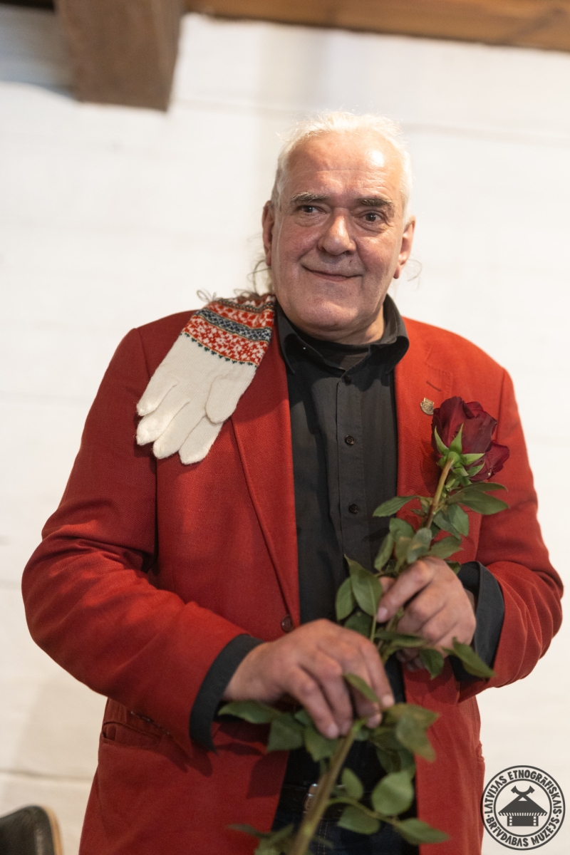 Smaidošs mākslinieks Jānis Žīmants; uz pleca rakstaini Rucavas pirkstaiņi, rokās sarkanas rozes. 