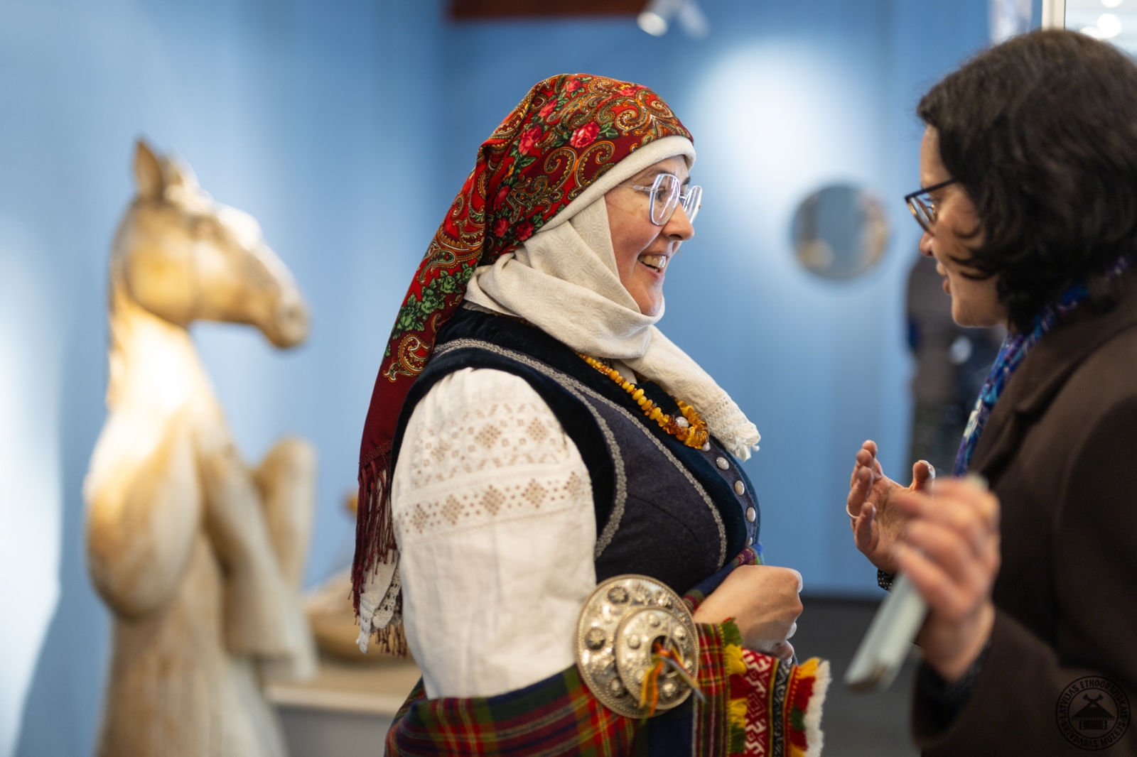 Divas sievietes sarunājas; viena tērpta greznā Rucavas tautastērpā, fonā koka zirga skulptūra. 