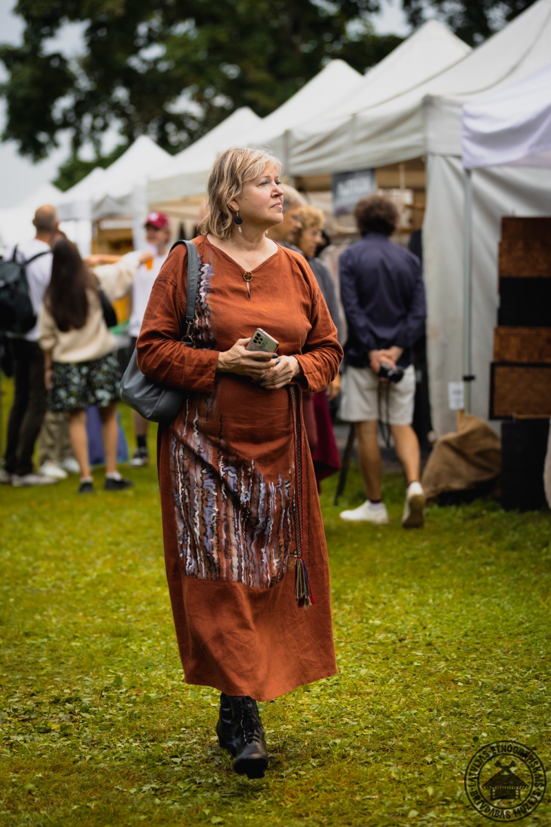 Tirgus apmeklētāja brūnā, stilīgā kleitā pēta amatnieku darbus. 
