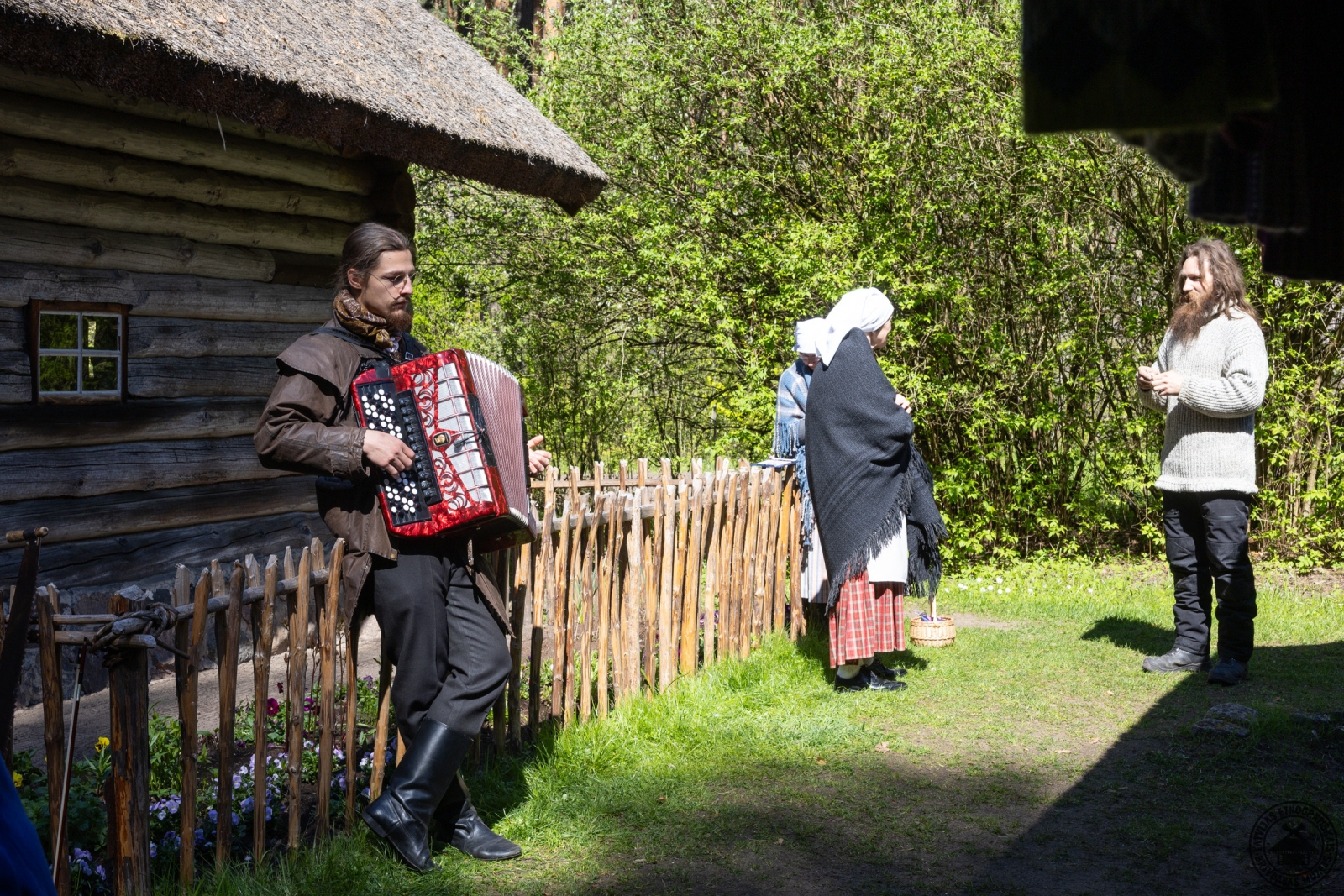 Akordeonists spēlē mūziku, fonā sarunājas trīs cilvēki tautastērpos. 