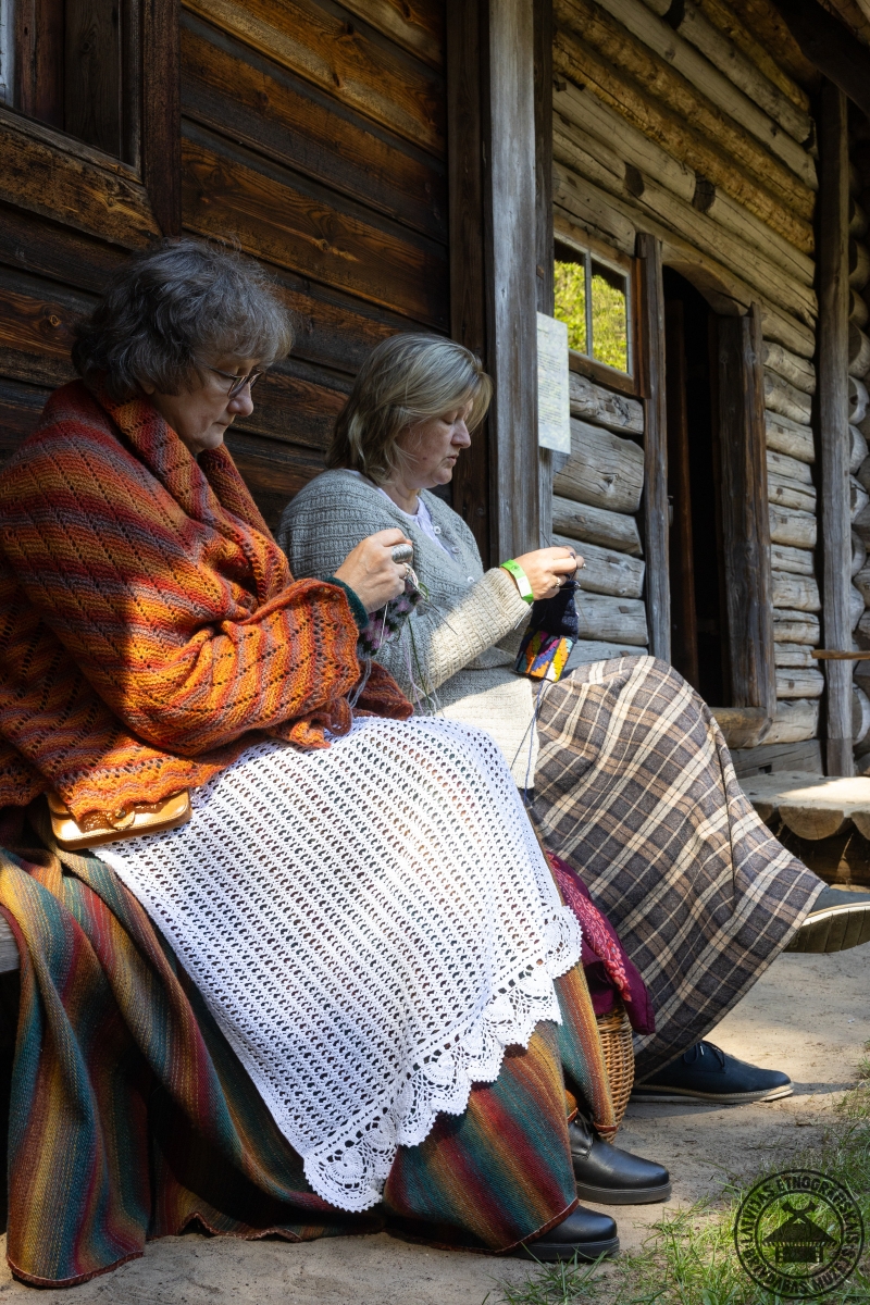 Divas sievietes tautastērpos sēž uz koka soliņa un kopīgi ada. 