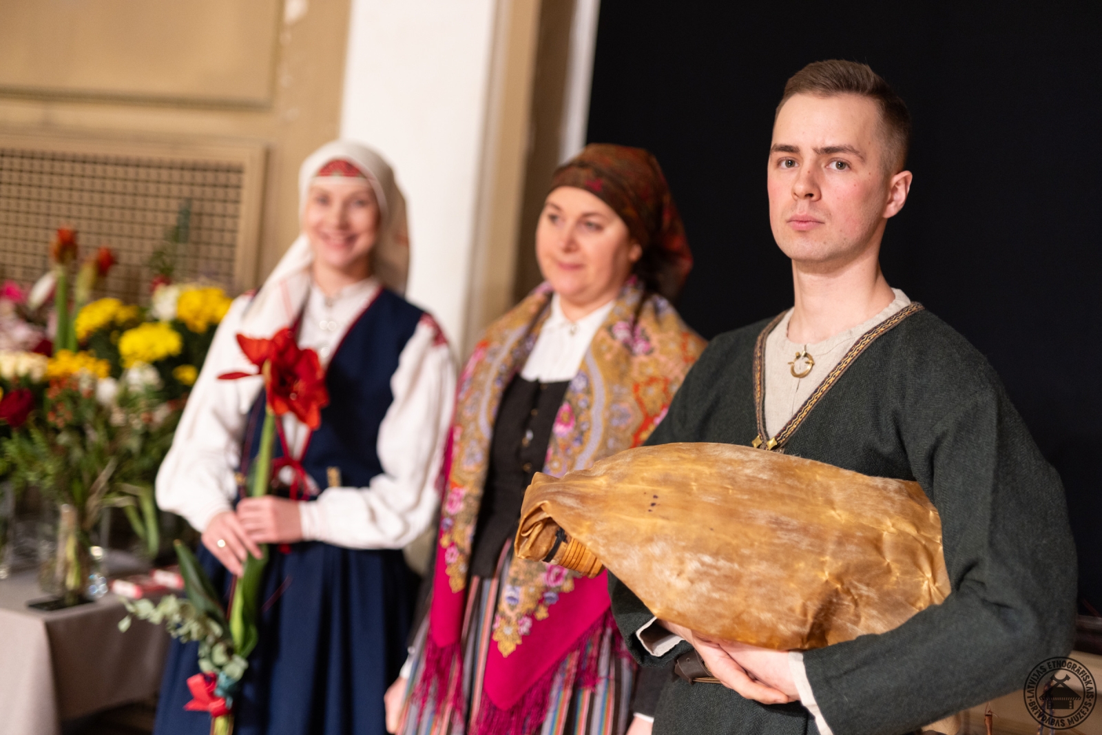 Kultūras ministrijas valsts sekretāre sveic muzeju simtgadē; priekšplānā darbinieki tautastērpos. 